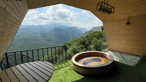 A wooden interior with a round hot tub, a table, and a view of a mountainous landscape. - Lost In Sky (Yenokavan)
