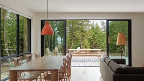Un interior moderno con una mesa y sillas de madera, un sofá y ventanas amplias que ofrecen vistas a un área boscosa.