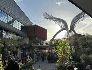 An outdoor shopping area with a modern building, a unique metal sculpture, and a variety of shops.