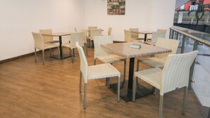 Un espace repas avec des chaises et des tables en osier, un ventilateur au mur et une vue sur les bâtiments à travers une rambarde en verre.