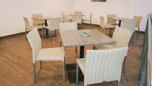 Un espace repas avec des chaises et des tables en osier, un menu bleu et un tableau représentant un paysage urbain accroché au mur.