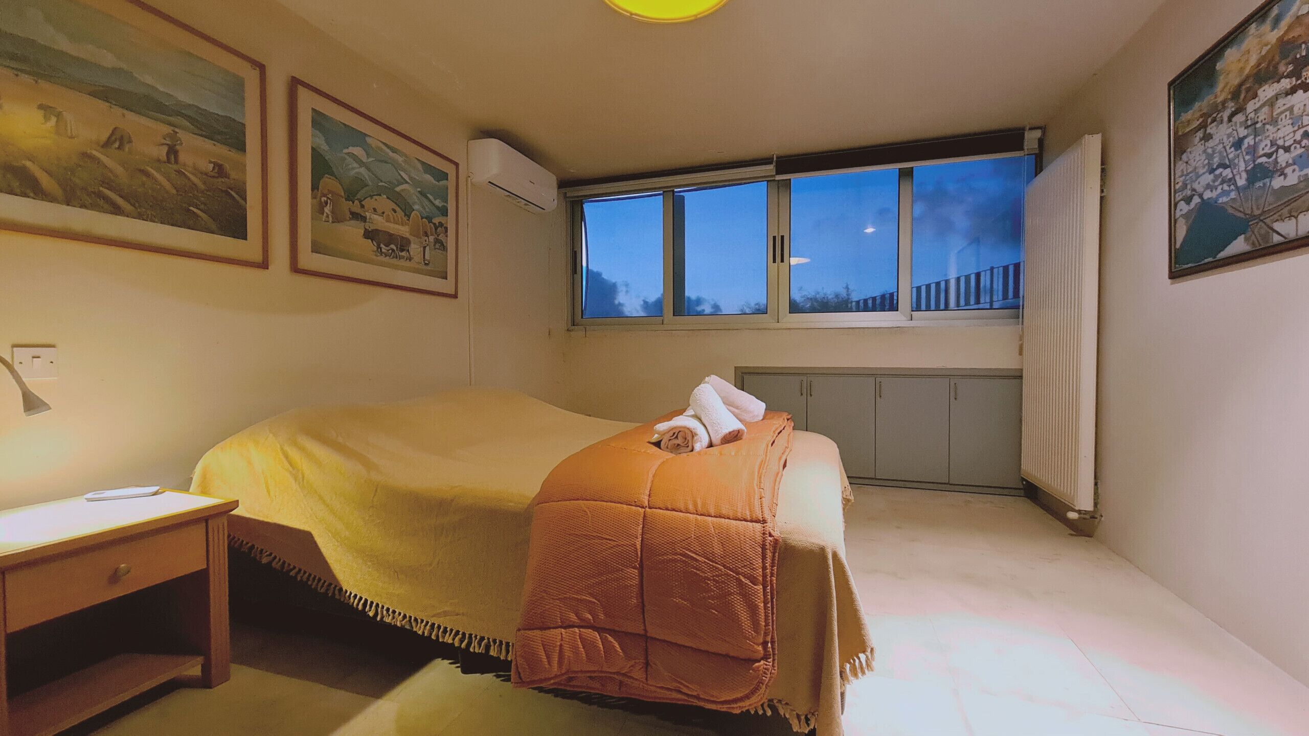 A bedroom with a bed, a nightstand, a window with a view, and two framed pictures on the wall.