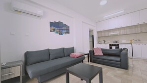 Un saló modern amb un sofà gris, una taula petita i una zona de cuina amb armaris i electrodomèstics blancs.