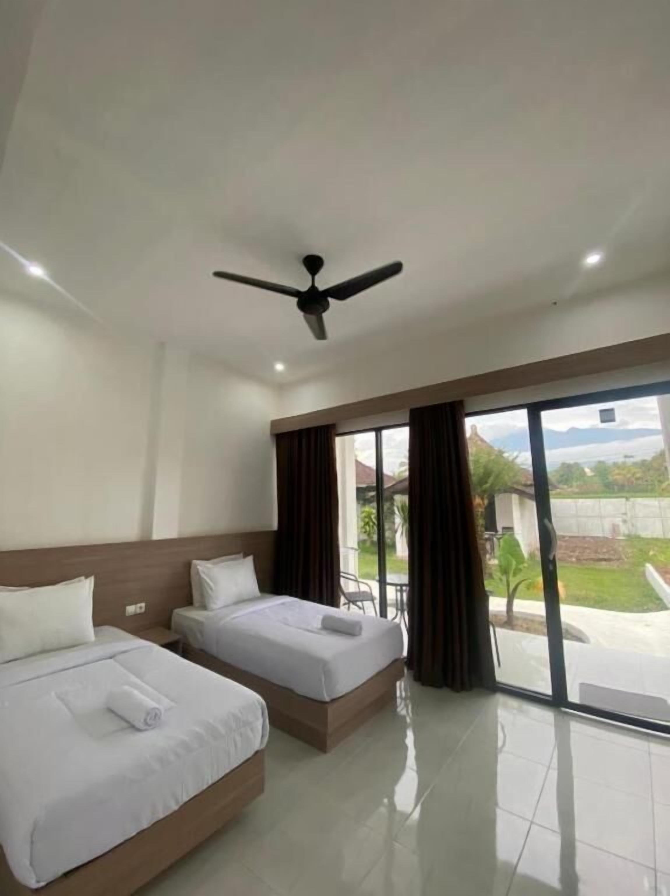 A hotel room with two beds, a ceiling fan, and a large window with a view of the outdoors.