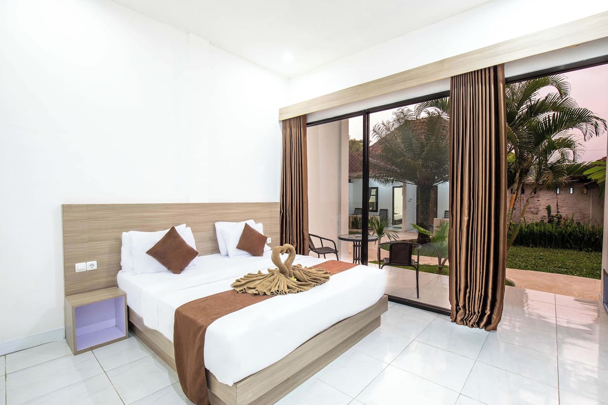 Habitación de hotel con una cama grande, una mesita de noche y un vidrio que da a un patio con mobiliario exterior.