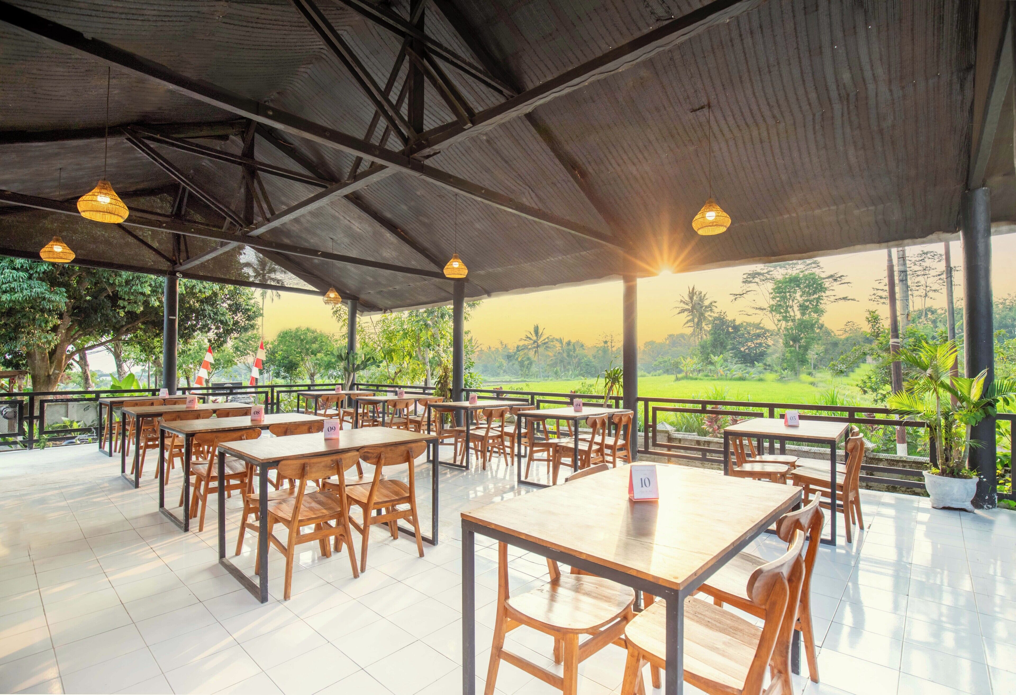 An outdoor dining area with wooden tables and chairs, a covered roof, and a view of a green landscape.