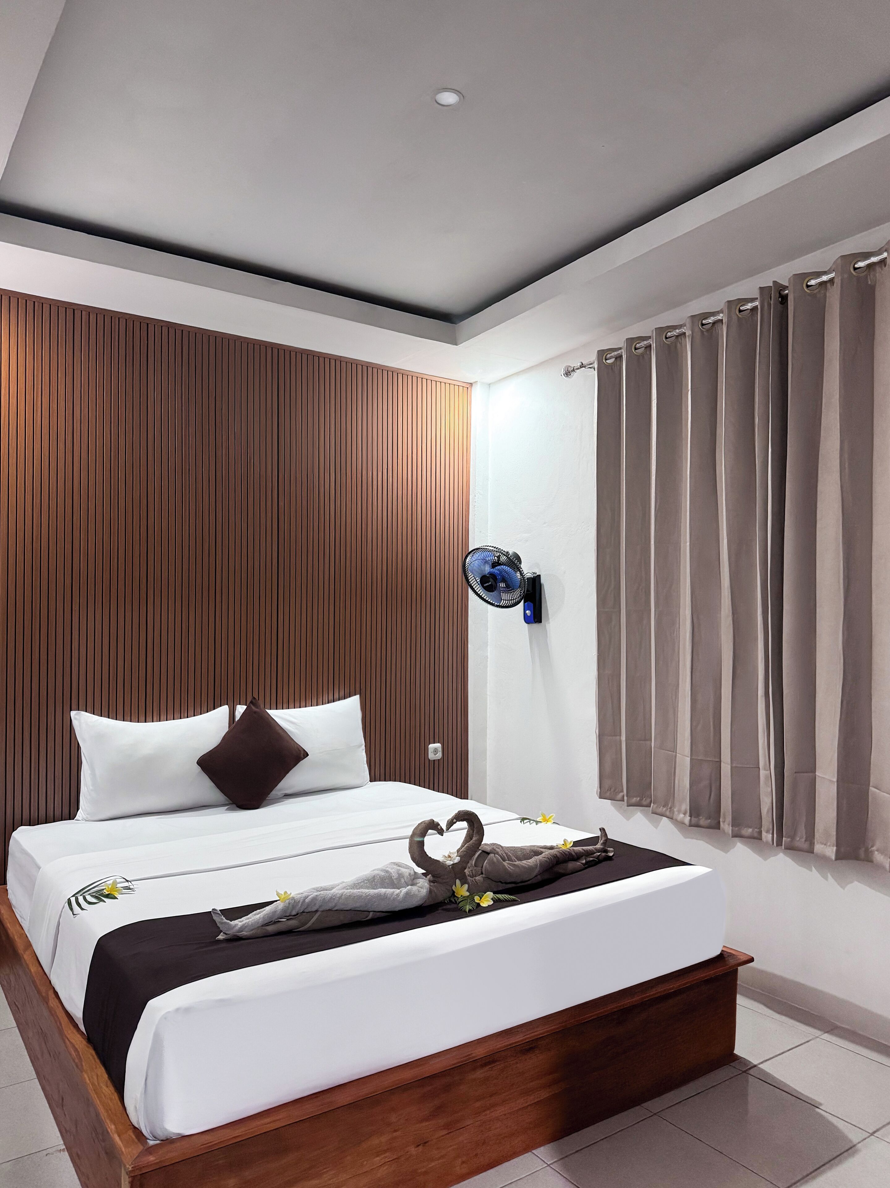 A modern hotel room with a wooden headboard, a bed with white bedding, a wall-mounted fan, and a window with curtains.