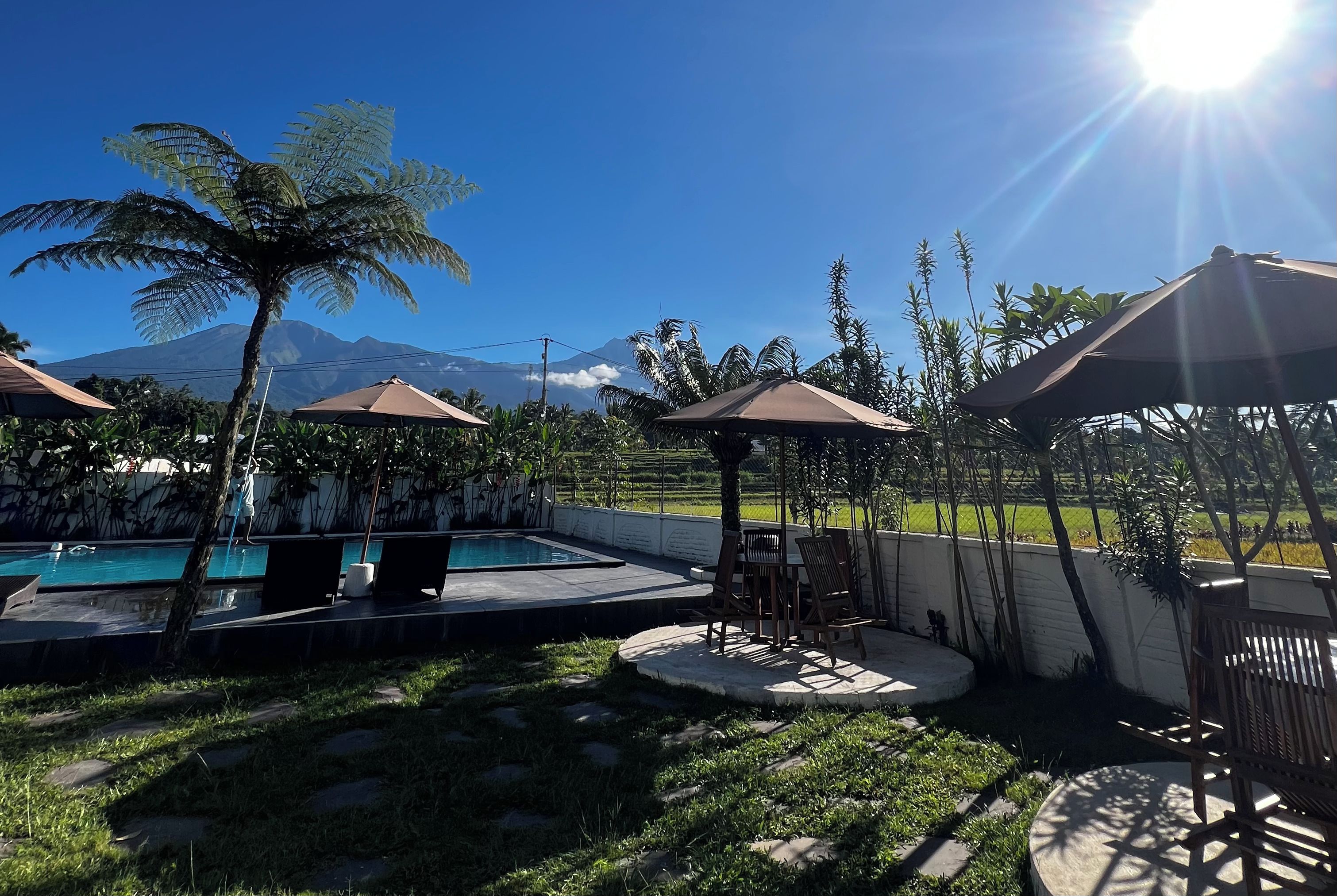 Zona de piscina con sombrillas, una mesa y sillas, rodeada de vegetación y vistas a la montaña.