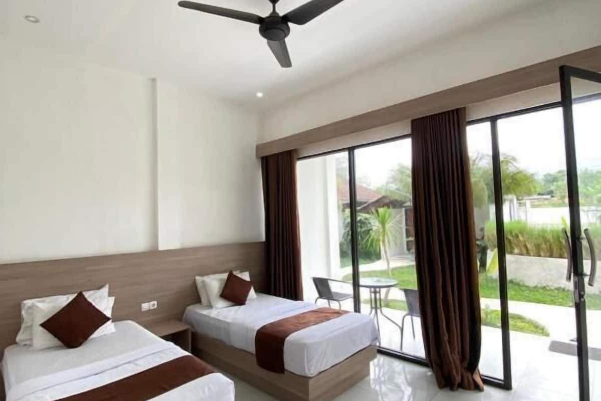 Habitación de hotel con dos camas, ventilador de techo y ventanas amplias con cortinas.