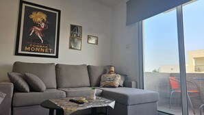 Un salon moderne avec un canapé gris, une petite table sur laquelle est posée une plante, et une grande fenêtre donnant sur l’extérieur.