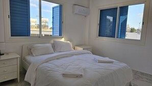 Une chambre à coucher avec un lit, du linge de lit blanc, une table de chevet, une lampe et deux fenêtres avec des volets bleus.