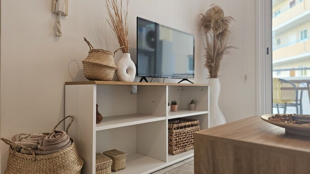 A living room with a TV on a wooden stand, wicker baskets, and a vase with dried plants. - STAY Miracle Apartment (Larnaca)
