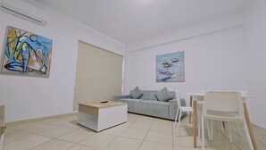 A small, modern living room with a sofa, a coffee table, and a dining table with chairs. There are two paintings on the walls.