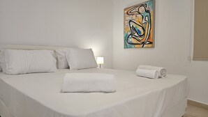 A neatly made bed with white linens and pillows, a rolled white towel on the bed, and a painting on the wall.