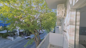 Un balcon donnant sur une rue bordée d’arbres, une voiture garée et un immeuble à la façade blanche.