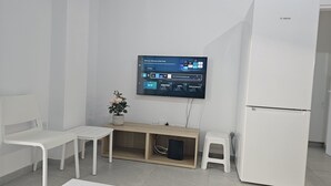 A modern, minimalistic room with a flat-screen TV mounted on the wall, a wooden desk with a computer, two white chairs, and a white refrigerator. - STAY Historia Apartment (Nicosia)