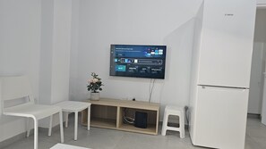 A modern, minimalistic room with a flat-screen TV mounted on the wall, a wooden desk with a computer, two white chairs, and a white refrigerator. - STAY Historia Apartment (Nicosia)