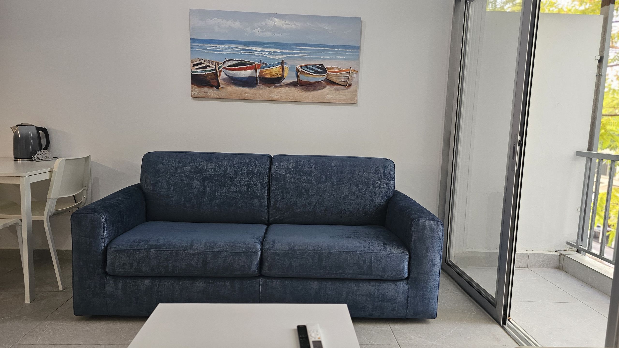 A living room with a blue sofa, a white coffee table, and a painting of boats on a beach.