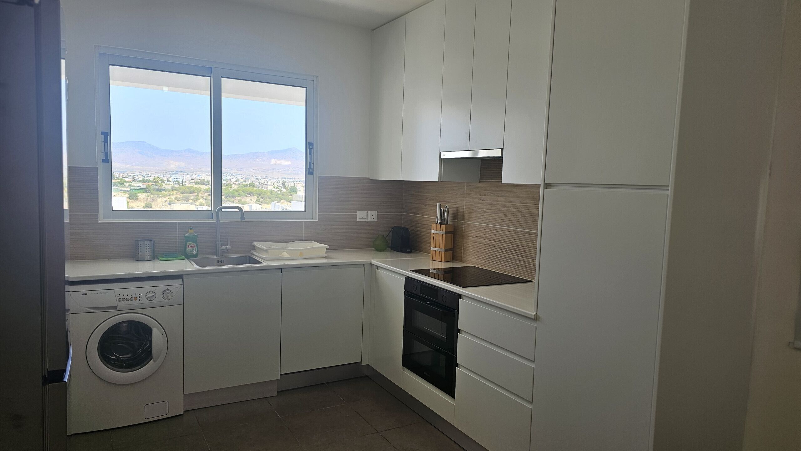 A modern kitchen with a built-in washer and dryer, a large window with a view, and a wooden backsplash.