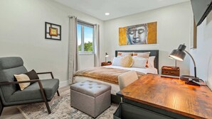 A bedroom with a bed, a chair, a desk, a lamp, a window with curtains, a painting on the wall, and a small table. - Good Vibes Sanctuary, Central Location South Miami (Cutler Bay)