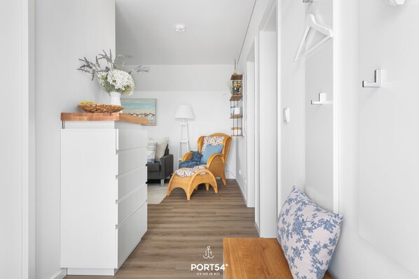 Un pasillo estrecho con un armario blanco, sillas de mimbre, un banco de madera y un estante de pared con objetos decorativos.