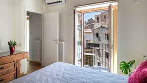 Een slaapkamer met een bed, een dressoir, een radiator, een airconditioner en een balkon met uitzicht op gebouwen.