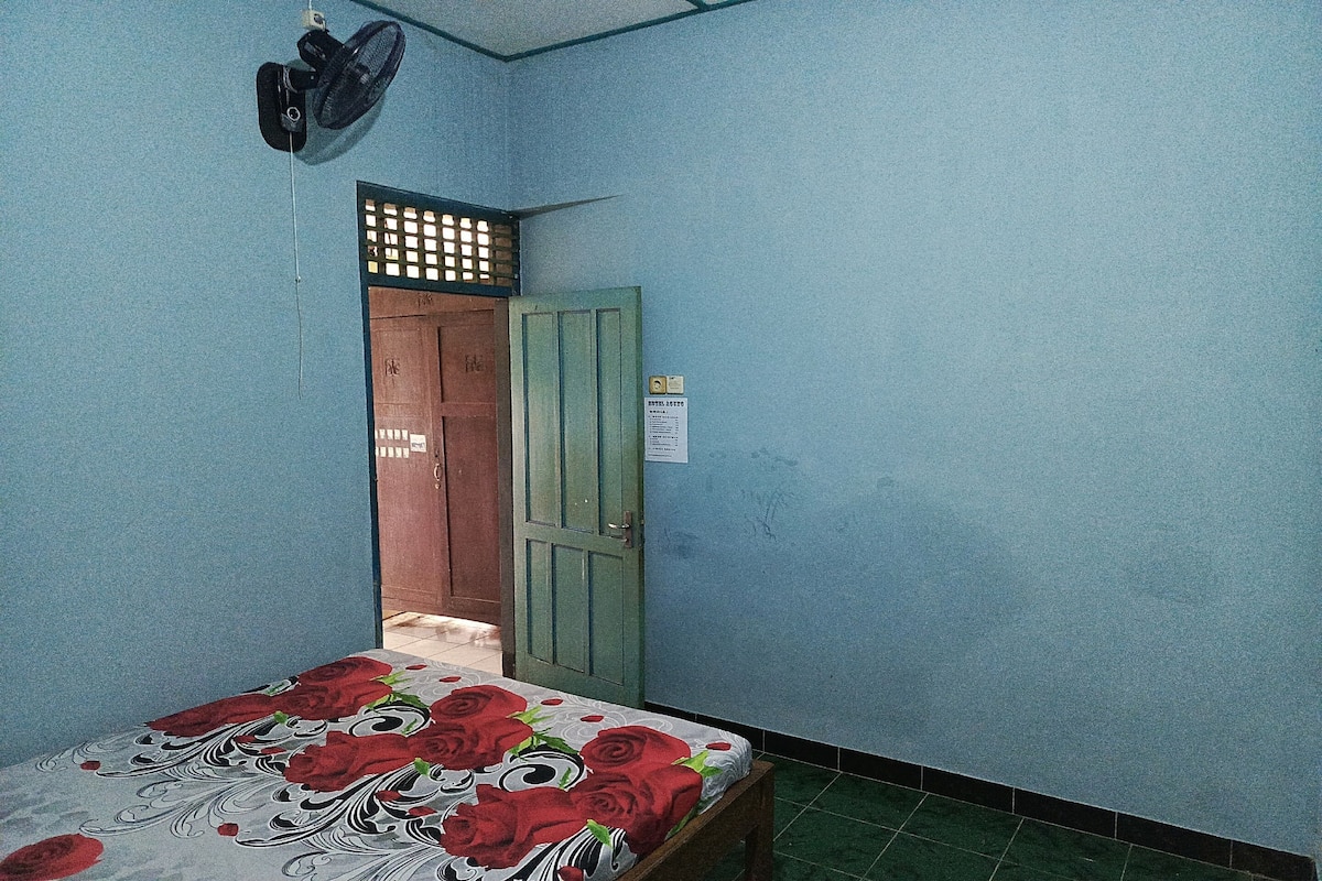 Um quarto com cama, porta e ventoinha fixada na parede.