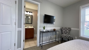 Habitación de hotel con cama, televisor en la pared, una mesita con una planta, una silla y un baño visible a través de una puerta abierta.