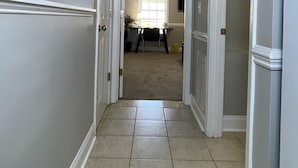 Un pasillo con detalles blancos, suelo de baldosas y una puerta que da a una habitación con ventana y escritorio.