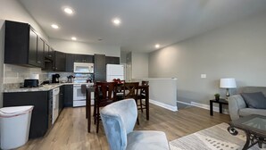 Una cocina moderna con armarios oscuros, un refrigerador blanco y un microondas. Hay una zona de comedor con sillas de madera y una mesa. En la sala de estar hay un sofá gris y una mesita con una lámpara.