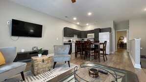 Una sala de estar moderna con televisor de pantalla plana, sillones grises, una mesa de centro y una zona de cocina con armarios oscuros y un refrigerador blanco.