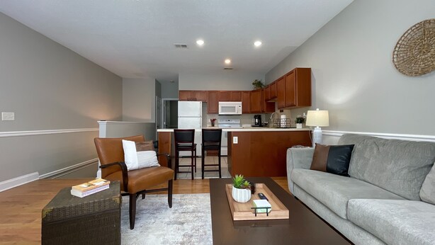 A modern living room with a gray sofa, two armchairs, a coffee table, and a small kitchen area with wooden cabinets and a microwave. - Elegant 2BR | Modern Amenities & Convenient Flair (Johnson City)