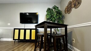 Un comedor con una mesa de madera y dos taburetes, una planta en maceta, un televisor montado en la pared y un adorno mural.