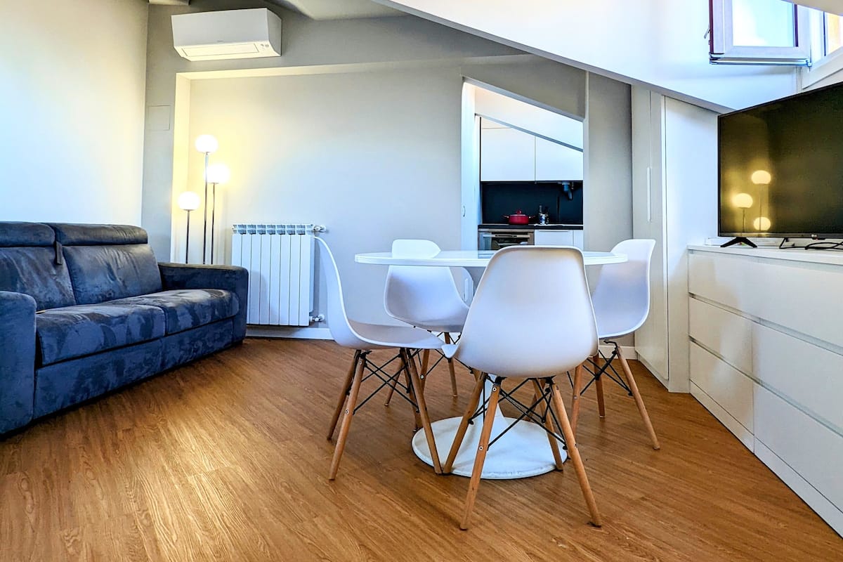 A modern living room with a sofa, dining table, and chairs, a kitchen area with a TV, and a large window.
