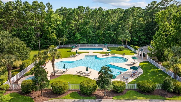A large, irregularly shaped swimming pool surrounded by a white fence, with multiple lounge chairs and umbrellas. - Spacious 3BR True Blue Condo | Pool Access • Close to Golf & Beach (Pawleys Island)