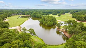 A golf course with a pond, surrounded by dense trees and a distant building. - Spacious 3BR True Blue Condo | Pool Access • Close to Golf & Beach (Pawleys Island)