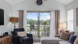 A living room with a black leather sofa, a grey sofa, a wooden cabinet, and a glass door leading to a balcony with a view of greenery. - Spacious 3BR True Blue Condo | Pool Access • Close to Golf & Beach (Pawleys Island)