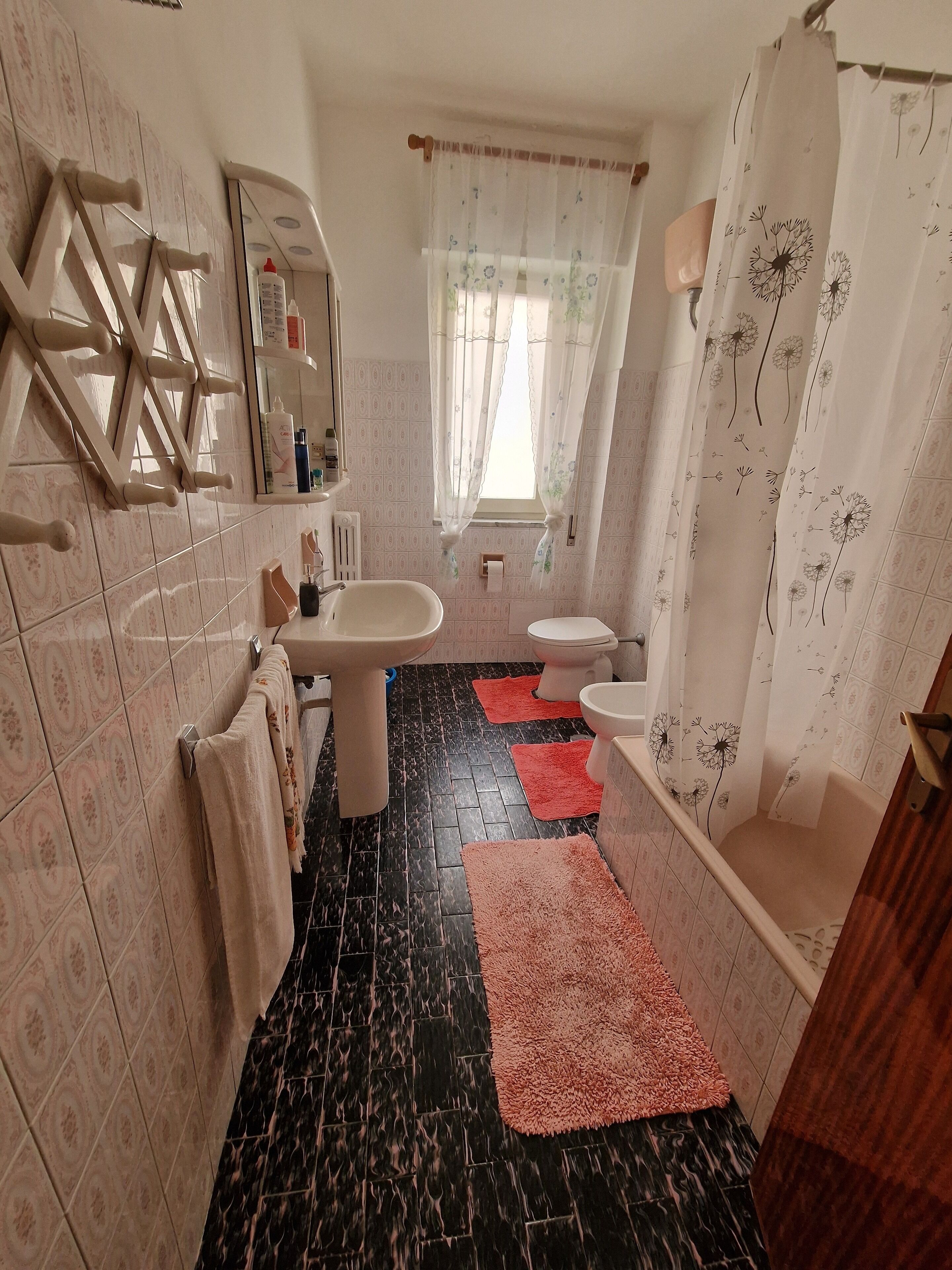 Een badkamer met een wastafel, toilet en bad. Handdoeken hangen aan de muur en er is een spiegel boven de wastafel. De vloer is voorzien van zwarte en roze vloerkleden.