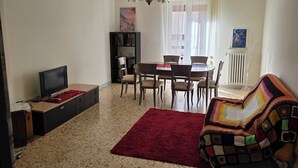 Un salon comprenant un téléviseur, une bibliothèque, une table à manger avec des chaises, une couverture colorée et un radiateur.