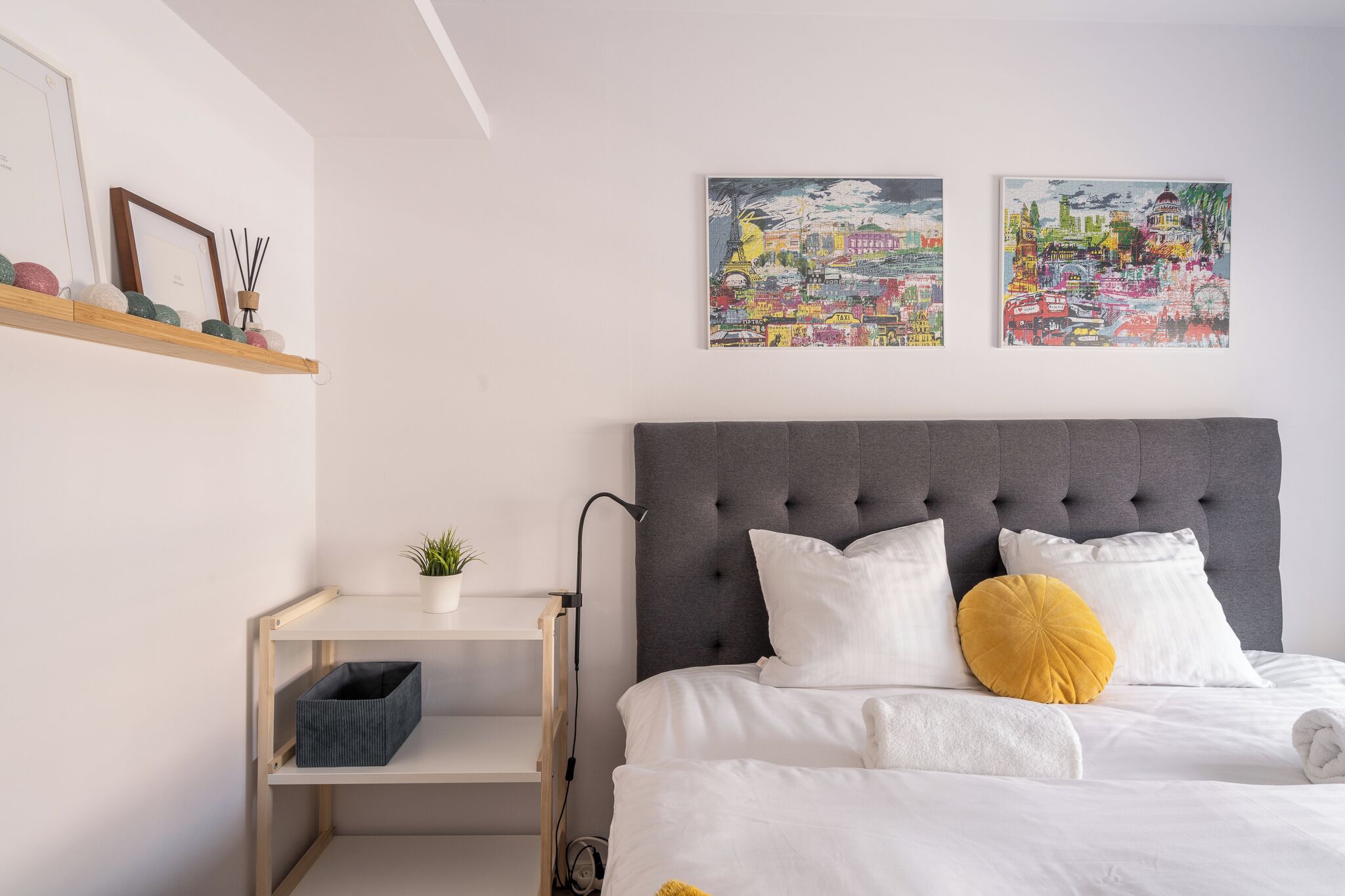 A neatly arranged bedroom with a grey tufted headboard, white bedding, and two colorful cityscape paintings on the wall.
