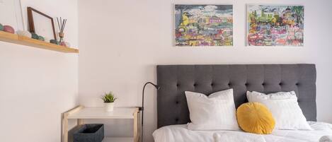 A neatly arranged bedroom with a grey tufted headboard, white bedding, and two colorful cityscape paintings on the wall.