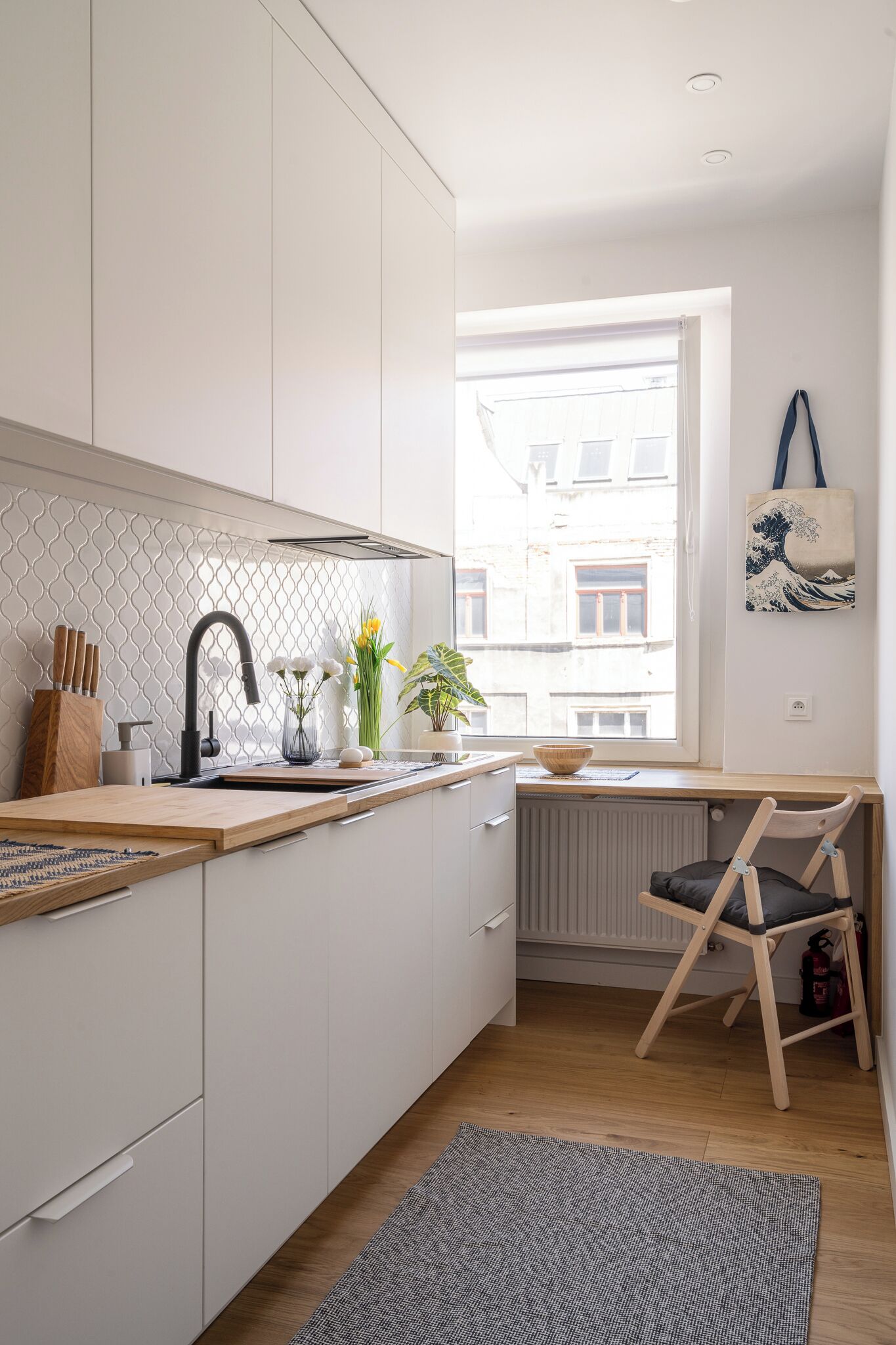 Cocina moderna con armarios blancos, un grifo negro y una encimera de madera. Se aprecia una ventana con vistas a edificios.