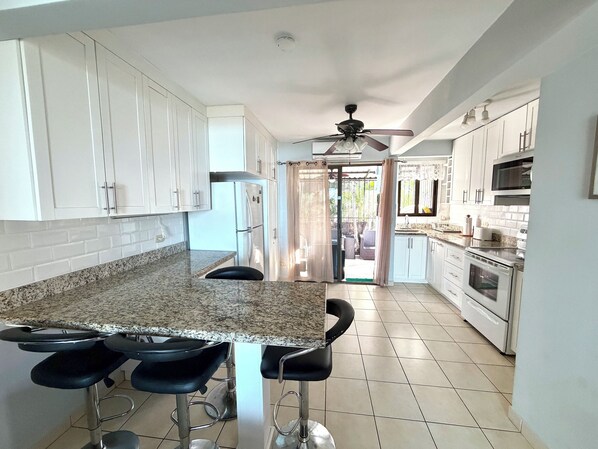 A modern kitchen with white cabinets, granite countertops, and a central island with bar stools. - Coco Maya 2b/2b Villa (Playas Del Coco)