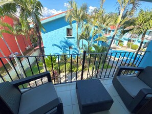 A balcony with a view of a swimming pool, surrounded by palm trees and a blue building.