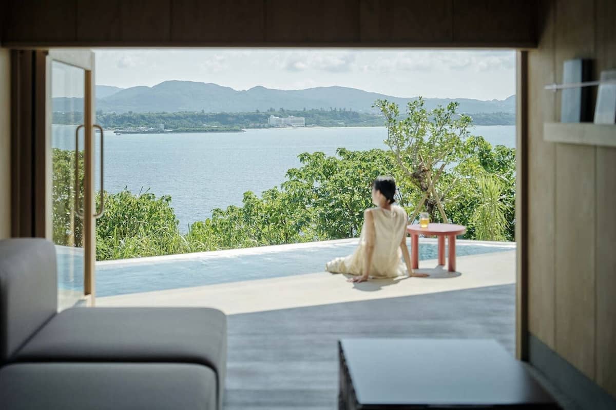 Una habitación con vista al agua, una persona sentada junto a la ventana y una mesita con una bebida.