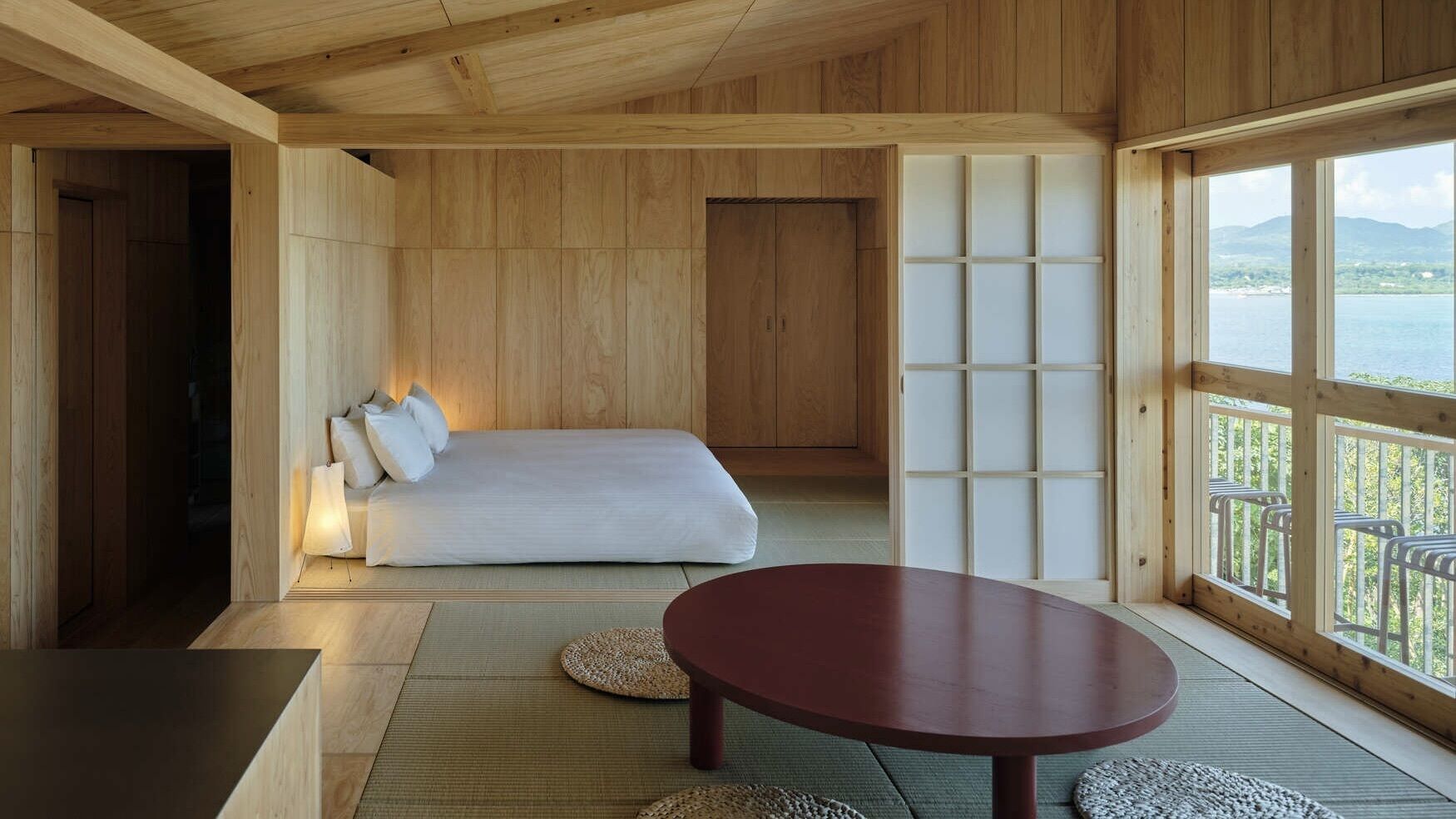 Ein Zimmer mit einem Bett, einem runden Tisch, Tatami-Matten und Blick auf ein Gewässer durch große Fenster.