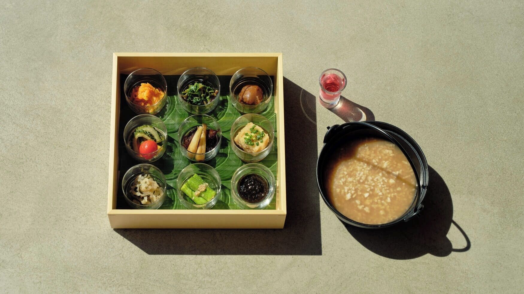 Une boîte en bois contenant neuf petits récipients en verre remplis de différents aliments, et un bol noir à côté duquel se trouve un liquide.
