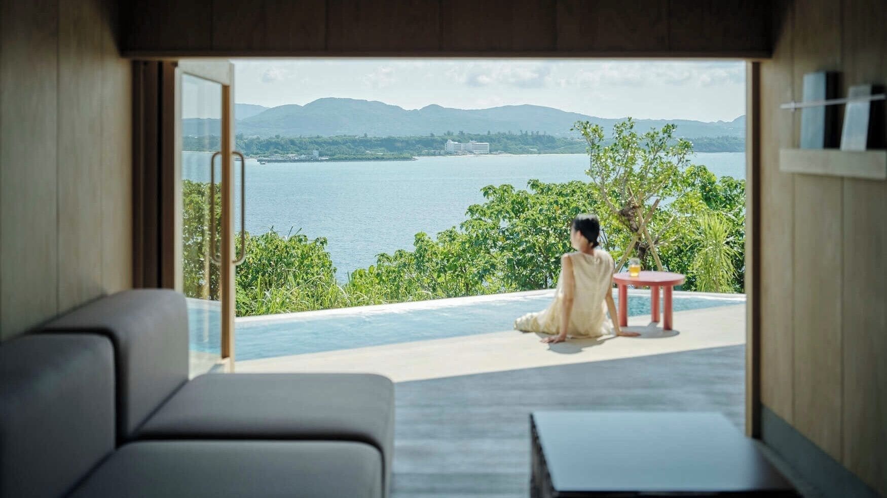 Una habitación con un amplio ventanal que da a una masa de agua y vegetación, una persona sentada junto a la ventana, y una mesita con una bebida.
