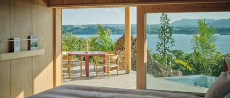 A bedroom with a bed, a wooden table, and chairs, and a view of a lake and mountains through large windows.