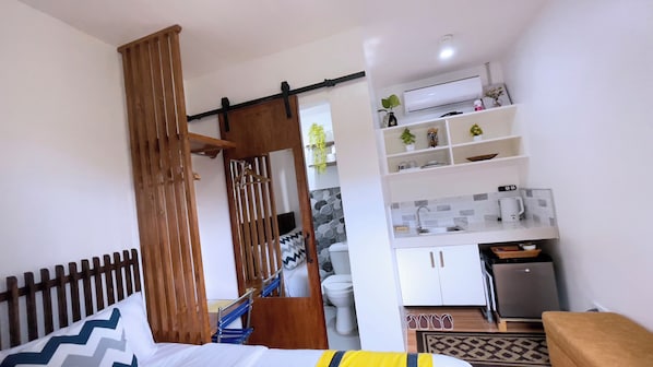 Habitación compacta con cama, un separador de madera, kitchenette con pileta y un refrigerador pequeño.